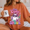 Woman sitting on a bed wearing a burnt orange t-shirt featuring the Too Cute to Spook graphic and holding a white ghost coffee mug.