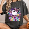 Smiling blonde woman sitting cross-legged wearing a washed dark gray t-shirt printed with the Feeling Wickedly Awesome witch ghost graphic, holding an orange jack-o-lantern mug